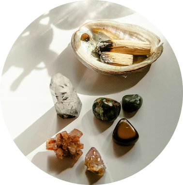 A top-down view of an abalone shell holding smoldering palo santo wood, surrounded by a variety of crystals including clear quartz and a reddish cluster, all arranged on a white circular surface with dappled sunlight shadows.